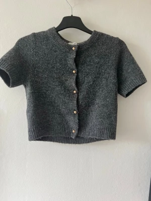 Mörkgrå kortärmad kofta från H&M - Kortärmad mörkgrå kofta med rund hals och pärlemorknappar. Koftan är stickad i ett mjukt material och har ribbade kanter vid ärmslut och nederkant. Perfekt att styla över en topp eller klänning för en trendig lager-på-lager-look.