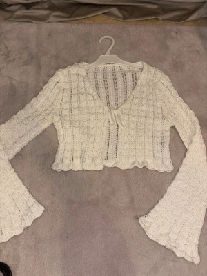 Vit stickad croppad tröja med trumpetärm - Supersöt vit stickad croppad tröja med v-ringning och knyt framtill. Tröjan har långa trumpetärmar och vågig kant nertill. Perfekt för dig som gillar en trendig och unik look. Materialet känns mjukt och stretchigt.