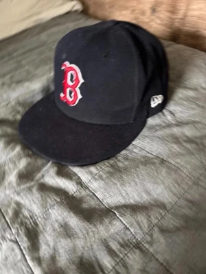 Svart Boston Red Sox keps New Era - Säljer en svart keps från New Era med Boston Red Sox-logga i rött och vitt framtill. Klassisk rak skärm och broderad logotyp på sidan. Perfekt för dig som gillar sportig stil och vill ha en ikonisk accessoar.