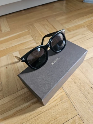 Svarta solglasögon från Tom Ford - Snygga svarta solglasögon från Tom Ford med klassisk rundad form och mörka glas. Bågarna är i blank svart plast med guldfärgade detaljer vid gångjärnen och logotyp på sidan. Perfekta för dig som vill ha en stilren och exklusiv look.