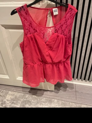 Rosa spetsdetaljerad topp Vero Moda - Snygg rosa topp från Vero Moda med spetsdetaljer upptill och omlott framtill. Toppen är ärmlös och har en lätt volang nedtill samt öppen rygg. Perfekt för dig som vill ha en feminin och trendig look.