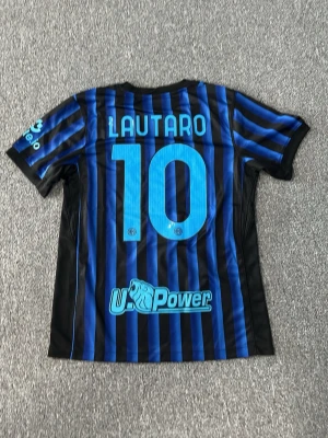 Inter Milan matchtröja Lautaro #10 - Snygg svart och blårandig Inter Milan matchtröja från Nike med Lautaro och nummer 10 på ryggen. Tröjan har klubbmärke, sponsorloggor och korta ärmar. Tillverkad i lätt och ventilerande material, perfekt för fotboll eller träning.