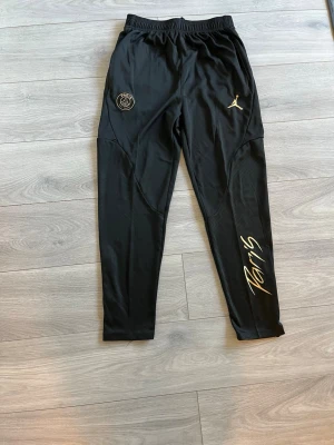 Svarta PSG x Jordan träningsbyxor - Snygga svarta träningsbyxor från Jordan i samarbete med PSG. Byxorna har guldfärgade detaljer, PSG-logga och Jumpman-logga framtill samt texten 'Paris' på ena benet. Tillverkade i ett lätt och stretchigt material med resår i midjan.