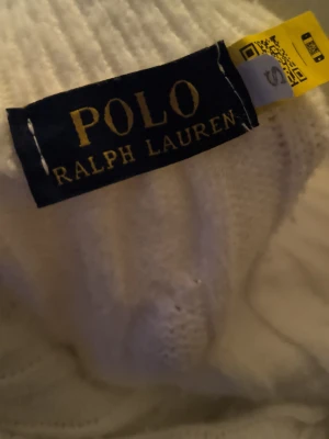 Vit kabelstickad tröja Polo Ralph Lauren - Vit kabelstickad tröja från Polo Ralph Lauren med halv dragkedja och broderad logga på bröstet. Tröjan har lång ärm, ribbade muddar och klassisk passform. Perfekt för en stilren och tidlös look.