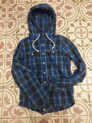 Blå hollister skjorta - Säljer denna feta blårutiga skjorta från Hollister med huva och vita snören. Den är i storlek M men sitter mer som S!! Dubbelkolla måtten!!  Hör av er vid frågor!!🤙🏻. Längd: 62 cm.    Bredd: Ca 42 cm
