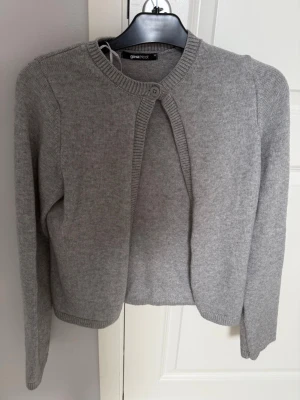 Grå kort kofta från Gina Tricot - Stilren grå kofta från Gina Tricot med rund halsringning och knapp framtill. Modellen är kortare och har långa ärmar. Materialet är mjukt och stickat, perfekt för lager på lager och enkel att matcha med olika outfits.