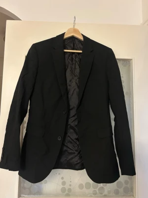 Svart klassisk kavaj med knappar - Stilren svart kavaj med två knappar framtill och fickor med lock. Kavajen har lång ärm och klassisk krage, perfekt för en dressad look. Fodrad insida för extra komfort. Passar till många olika outfits.