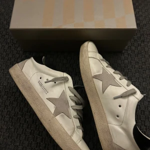 Golden Goose vita sneakers med stjärna - Golden Goose sneakers i vitt skinn med grå mockadetaljer och ikonisk stjärna på sidan. Skorna har grå snörning, rund tå och platt sula. Klassisk streetstil med perforerade detaljer på plösen och svart hälparti. Säljes med originalkartong. Lätt skada på vänster häl