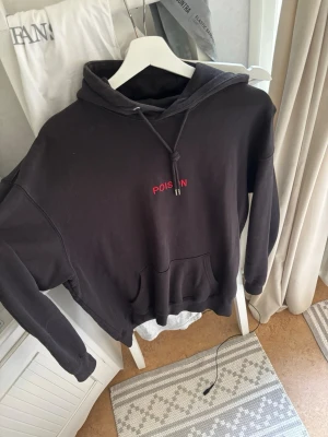 Svart hoodie med röd POISON-tryck - Cool svart hoodie med röd POISON-text och ormtryck på ryggen samt mindre POISON-tryck på bröstet. Klassisk känguruficka och dragsko i huvan. Perfekt för dig som gillar streetwear och vill sticka ut med en unik design.