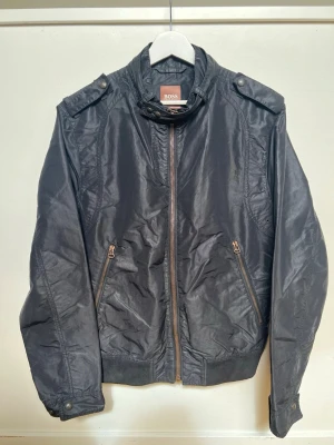 Svart vindjacka från BOSS - Stilren svart vindjacka från Hugo BOSS militär stil, med hög krage, dragkedja framtill och två snedställda fickor med dragkedja. Jackan har axelklaffar, ribbade muddar och är tillverkad i ett glansigt syntetmaterial. Perfekt för en cool och sportig look.