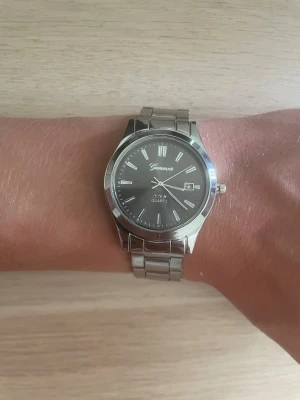 Silverfärgad klocka - Stilren klocka med silverfärgat armband i metall och svart urtavla. Klockan har romerska siffror, datumvisning och klassisk design. Perfekt accessoar för dig som gillar en clean och tidlös look. Liten skråma på baksidan av klockan. 