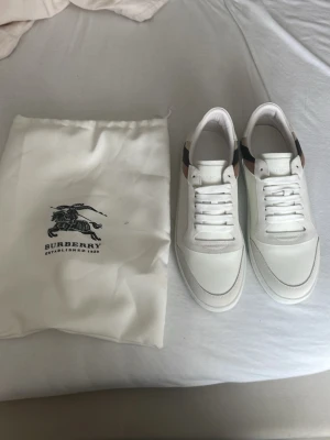 Vita Burberry sneakers med rutigt mönster - Snygga vita sneakers från Burberry med klassiskt beige, svart och brunt rutmönster på hälen. Skorna har vita snören, rund tå och är tillverkade i läder och textil. Perfekta för dig som vill ha en stilren och exklusiv look.