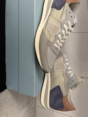 Philippe Model grå sneakers - Snygga sneakers från Philippe Model i olika nyanser av grått, beige och blått. Ovandelen är i en mix av mocka, mesh och läderdetaljer. Klassisk låg modell med vita skosnören och kontrastfärgad häl i brunt. Perfekta för en clean och stilren look. Aldrig använda