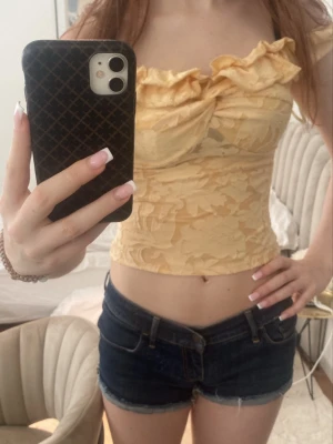 Gul croppad topp med volang - Söt gul croppad topp med volangdetaljer och offshoulder-design. Toppen har ett subtilt blommönster och är figurnära med en romantisk vibe. Perfekt för varma dagar och somriga outfits.