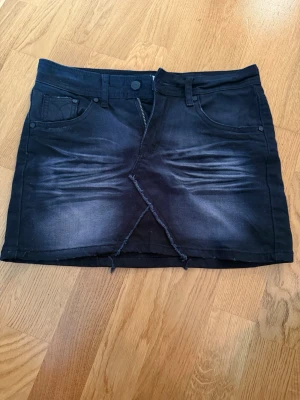 Svart jeanskjol med broderi och slits - Cool svart jeanskjol med slitningar och fransig slits både fram och bak. Kjolen har fem fickor, dragkedja och knäppning framtill samt snygga broderade detaljer bak. Perfekt för dig som gillar en edgy streetstil.