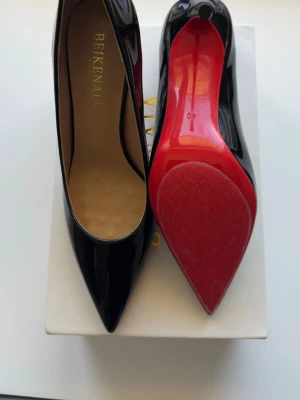 Svarta lackpumps med röd sula - Snygga svarta pumps i lack från Bekenaia med spetsig tå och hög klack. Skorna har en ikonisk röd sula och elegant siluett. Perfekta för dig som vill sticka ut med en klassisk men edgy look. Materialet är glansigt syntetläder.