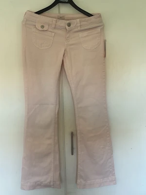 Ljusrosa bootcut jeans med fickor - Säljer ett par ljusrosa jeans med bootcut-modell och två stora fickor framtill med knappdetaljer. Jeansen har även bakfickor och bälteshällor. Perfekta för dig som vill ha en unik och trendig look. Materialet känns mjukt och stretchigt.