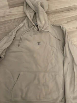 Beige hoodie från Les Deux - Säljer en stilren beige hoodie från Les Deux med broderad romersk siffra II på bröstet. Hoodien har huva med dragsko och en stor magficka. Perfekt för dig som gillar minimalistisk design och mjuka material.
