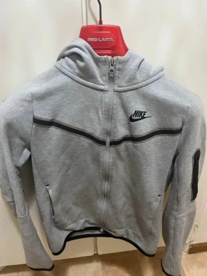 Nike tech - Snygg grå hoodie från Nike med dragkedja framtill och svart Nike-logga på bröstet. Hoodien har svarta detaljer längs bröstet och ärmarna samt en praktisk ficka på ena ärmen. Tillverkad i mjukt sweatshirtmaterial och har huva för extra komfort.