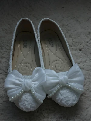 Vita ballerinaskor med rosett och pärlor - Supersöta vita ballerinaskor från GUOCOO med stor rosett framtill och dekorativa pärlor. Skorna har en texturerad yta och är täckta i tyg som ger en elegant look. Perfekta för dig som vill ha något extra till outfiten! STORLEK 39!!!