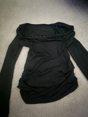 Svart off shoulder topp med nitar - Trendig svart off shoulder topp med långa ärmar och små silverfärgade nitar längs den vida urringningen. Tunn och stretchig kvalitet som sitter snyggt på kroppen. Perfekt för dig som gillar en edgy och stilren look.