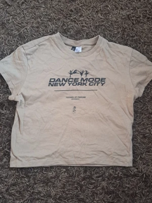 Beige croppad t-shirt - Beige croppad t-shirt från Dance Mode New York City med svart tryck framtill. Tillverkad i mjuk bomull, perfekt för en avslappnad streetstyle-look.