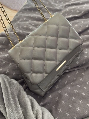 Grå quiltad handväska med kedjerem - Snygg grå handväska i quiltat konstläder med guldfärgad kedjerem och elegant metallspänne. Väskan har flera fack inuti för bra organisering och en stilren design som passar till många outfits.