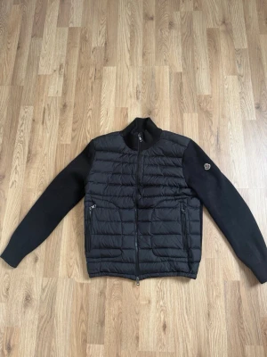 Svart cardiganjacka från Moncler - Snygg svart cardiganjacka från Moncler med quiltad front och stickade ärmar. Jackan har två dragkedjeförsedda fickor och en hög krage. Märke på ärmen och diskret logga på insidan. Perfekt för lager-på-lager och streetstyle.