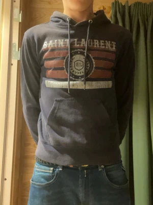 Mörkblå Saint Laurent hoodie - Säljer en mörkblå hoodie från Saint Laurent med tryck framtill i vitt och rött samt ett stort SL-emblem. Hoodien har huva och en stor magficka. Perfekt för dig som vill ha en snygg och bekväm tröja.