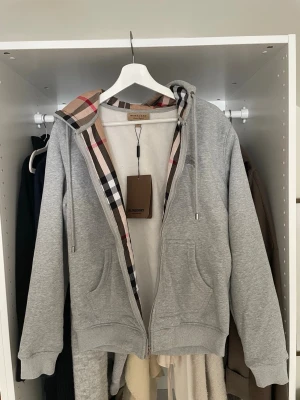 Grå zip hoodie (inte Burberry) - Säljer en grå hoodie med dragkedja och klassiskt rutigt foder i huvan. Hoodien har två fickor framtill, justerbar dragsko i huvan och ribbade muddar. Perfekt för dig som vill ha en stilren och lyxig vardagslook.