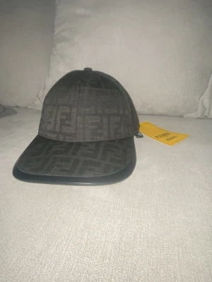 Svart keps från Fendi med logomönster     Jag har tagit av den gula lappen  - Snygg svart keps från Fendi med det klassiska FF-logomönstret över hela kepsen. Skärmen har en stilren svart kant i läderimitation och kepsen har en justerbar rem baktill. Perfekt accessoar för att lyfta din streetwear-look.