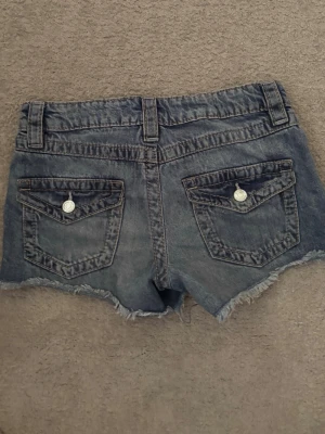 Blå jeansshorts med knappficka strl 152 från Gina Tricot  - Säljer ett par blå jeansshorts med fransig kant och slitna detaljer. Shortsen har fickor både fram och bak med knappdetaljer och bälteshällor. Perfekta för sommaren och ger en avslappnad look.