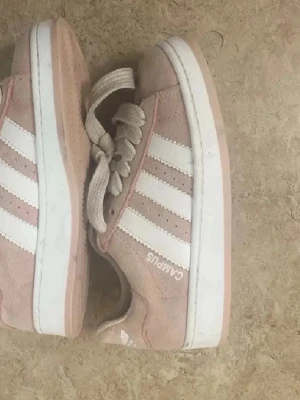 Adidas Campus rosa sneakers - Säljer ett par rosa Adidas Campus sneakers i mocka med klassiska vita ränder på sidorna och vit sula. Skorna har låga skaft och vita skosnören. Perfekta för dig som gillar retrostil och streetwear.