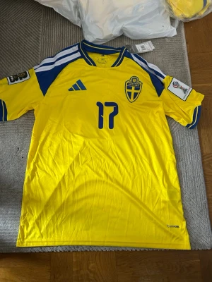 Svensk landslagströja Adidas gul/blå - Gul svensk landslagströja från Adidas med blå detaljer och nummer 17 på bröstet. Tröjan har officiellt Sverige-emblem, blåa ränder på axlarna och tryck på ärmarna. Tillverkad i lätt och ventilerande polyester, perfekt för fotboll eller träning.