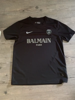 Svart PSG x Balmain tröja Nike - Snygg svart t-shirt från Nike i samarbete med PSG och Balmain. Tröjan har vita tryck med Balmain Paris-logga på bröstet, PSG-märke och Nike swoosh. Tillverkad i Dri-FIT material som andas, perfekt för träning eller sport. Pris kan diskuteras 