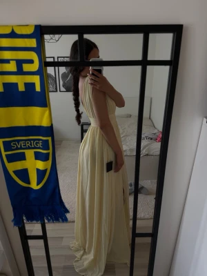 Ljusgul långklänning med v-ringning - Säljer en elegant ljusgul långklänning med djup v-ringning och breda axelband. Klänningen har ett markerat midjeband och ett luftigt, svepande fall. Perfekt för sommarens alla tillfällen.