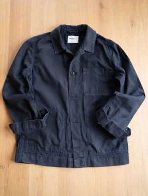 These Glory Days Chore Jacket, size M - Stilren svart overshirt från THESE GLORY DAYS i kraftig bomull. Klassisk rak passform med tre stora fickor framtill och knäppning med fyra knappar. Perfekt lager-på-lager-plagg med enkel krage och diskret design.