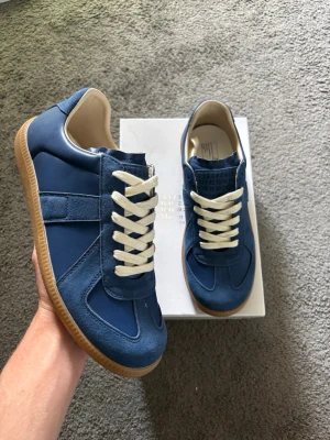 Blå Maison Margiela sneakers - Snygga blå sneakers från Maison Margiela med ovandel i mocka och läder, vita skosnören och klassisk gummisula. Ikonisk design med diskreta detaljer och låg profil som ger en clean och stilren look.