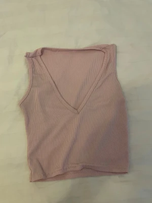 Ljusrosa ribbad linnetopp - Säljer en ljusrosa ribbad linnetopp med djup v-ringning. Toppen är ärmlös och har en slim passform som sitter snyggt på kroppen. Perfekt att matcha med jeans eller kjol för en trendig look.