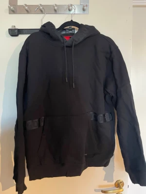 Svart hoodie med spännen - Svart hoodie med huva och dragsnören. Framtill finns två dekorativa spännen vid fickan som ger en unik look. Hoodien har klassisk känguruficka och ribbade muddar vid ärmslut och nederkant. Perfekt för dig som gillar streetwear och enkel stil.