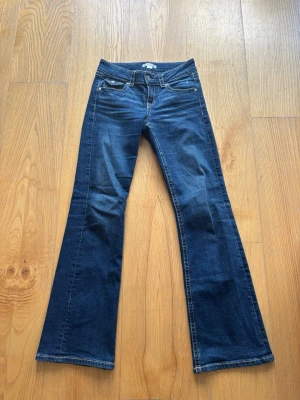 Mörkblå bootcut jeans - Säljer ett par mörkblå bootcut jeans med klassisk femficksdesign och kontrastsömmar. Jeansen har låg midja och utsvängda ben, perfekta för en retroinspirerad look. Materialet är mjukt denim som ger en bekväm passform.