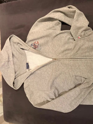 Grå zip hoodie från Champion  - Säljer en grå zip hoodie från Champion i storlek. Hoodien har broderad logga på bröstet och liten Champion-detalj på ärmen. Tillverkad i mjuk bomullsblandning med borstad insida för extra komfort. 