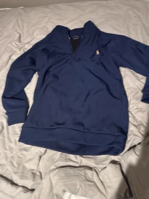 Marinblå tröja från Polo Ralph Lauren - Snygg marinblå långärmad tröja från Polo Ralph Lauren med dragkedja vid halsen och klassiska broderade loggan på bröstet. Tillverkad i mjukt material och har ribbade muddar vid ärmslut och nederkant. Perfekt för en stilren och avslappnad look.