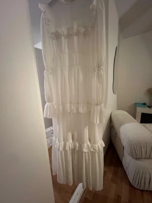 Vit långklänning med volanger - Långklänning i vit färg med flera lager av volanger och hög hals. Klänningen har långärmade ärmar och en luftig, romantisk design som ger ett drömmigt intryck. Perfekt för dig som gillar en bohemisk och elegant stil.