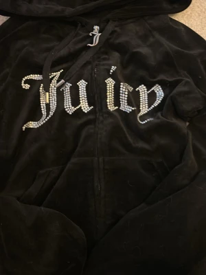 Juicy Couture svart velour hoodie - Snygg svart hoodie i mjuk velour från Juicy Couture med glittrande stenar som stavar 'Juicy' på bröstet. Dragkedja framtill, huva och fickor på sidorna. Perfekt för en chill och trendig look.