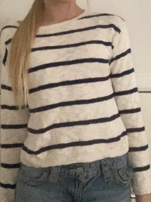 Vit stickad tröja med blå ränder - Säljer en vit stickad tröja med horisontella blå ränder. Tröjan har rund halsringning och långa ärmar. Perfekt till jeans för en avslappnad look. Materialet känns mjukt och lätt, vilket gör den skön att bära.