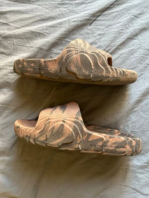 Beige camo tofflor från Adidas - Säljer ett par chunky tofflor från Adidas i beige och grått camomönster. De har en bred rem över foten och en riktigt tjock, räfflad sula för extra komfort. Tillverkade i ett mjukt, flexibelt material som känns skönt att gå i. Perfekta för chill dagar hemma eller på stranden.