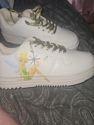 Vita sneakers med Tingeling och glitter - Unika vita sneakers med motiv av Tingeling på sidan och glittriga guldfärgade skosnören. Skorna har en klassisk låg modell med perforerade detaljer framtill och tjock sula. Materialet är syntetläder och designen är lekfull och trendig. Från Zalando Disney, ny pris ca 600kr 