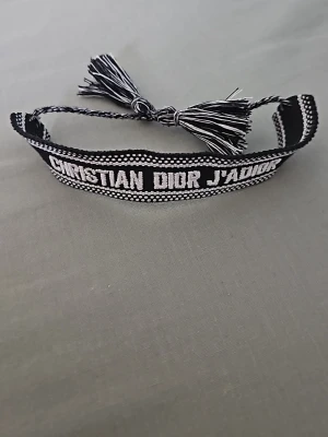 Christian Dior armband - Snyggt svart och vitt vävt armband från Christian Dior med texten 'J'adior'. Armbandet har knytning med två tofsar och är tillverkat i textil. Perfekt för att ge din outfit en exklusiv touch.
