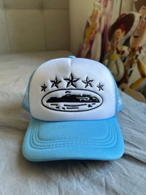 Blå och vit truckerkeps med stjärnor - Cool truckerkeps i blått och vitt med mesh på sidorna och bak. Framsidan har ett broderat UFO-motiv med fem stjärnor ovanför. Skärmen är böjd och kepsen har justerbar snapback. Perfekt för dig som gillar unika accessoarer och streetstyle.